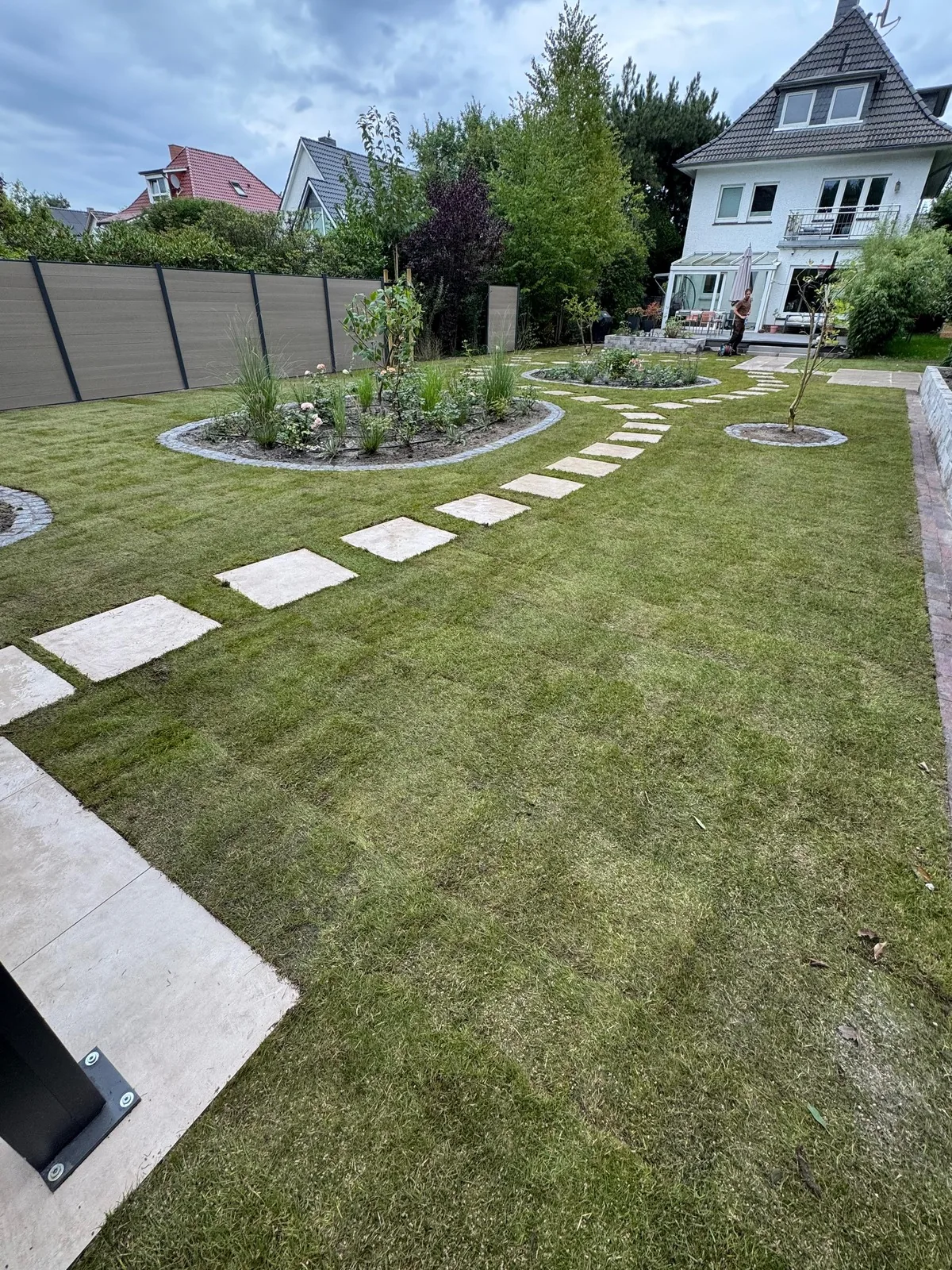 Fertiger Garten mit Blick zum Haus: Rollrasen, Trittplatten, zwei bepflanzte Rondelle mit Granitkante, links der Sichtschutz