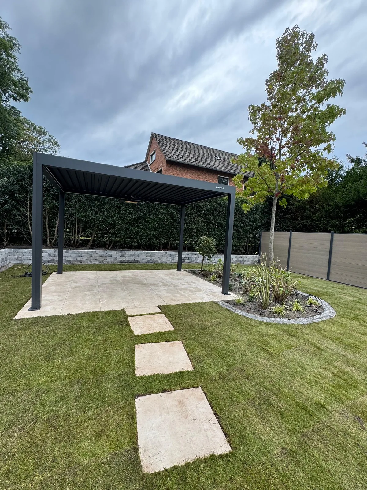 Pergola auf der fertigen Terrassenfläche, davor Trittplatten im Rollrasen, rechts ein bepflanztes Beet mit Granitkante