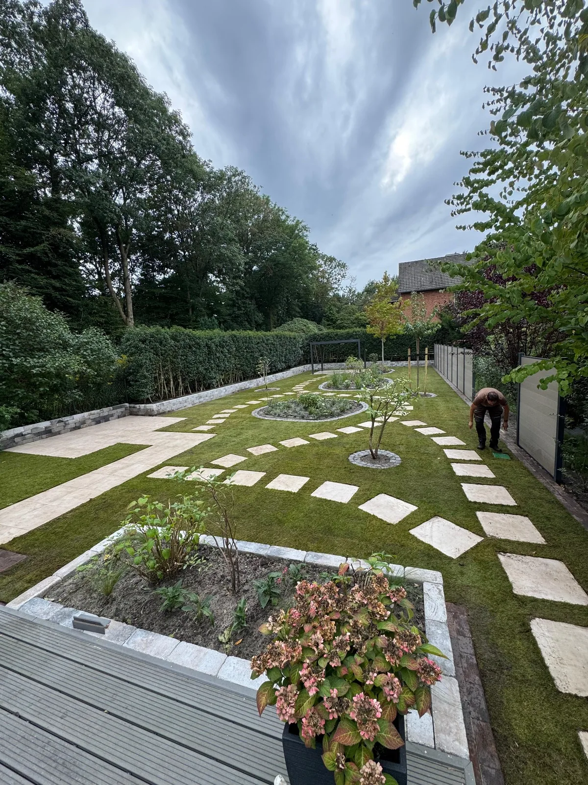 Blick von der Holzterrasse über den fertigen Garten: bepflanztes Hochbeet, Rollrasen mit Trittplatten, Rondell und Pergola im Hintergrund