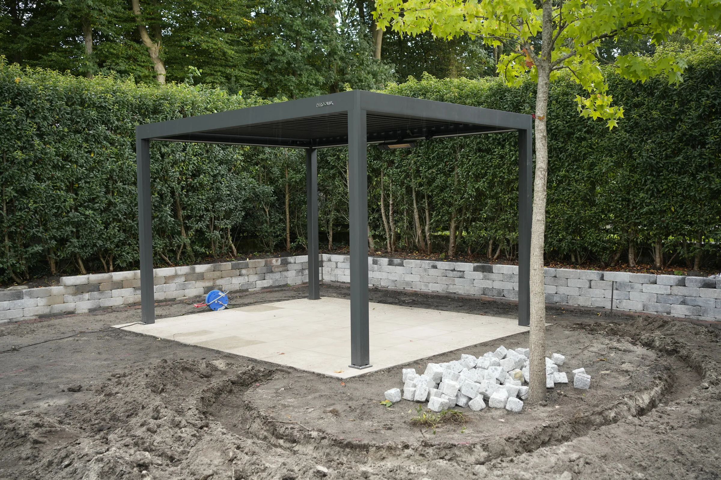 Neu gebaute Pergola auf einer hellen Terrassenfläche, eingefasst von einer Blocksteinmauer, die Beete rundherum noch offen