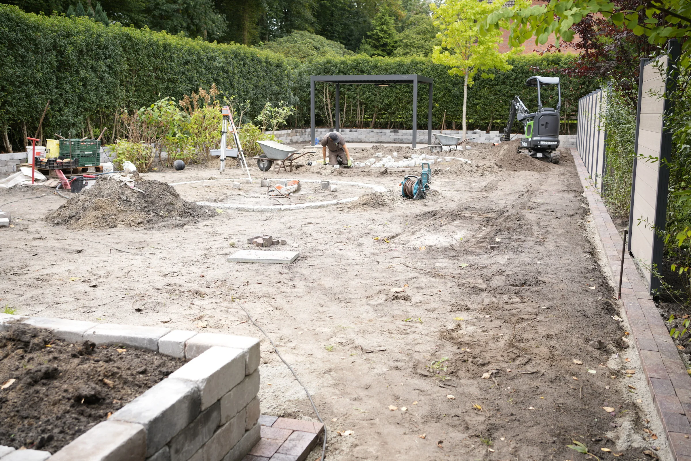 Gartenbaustelle im Rohbau: abgetragene Fläche, Blocksteinmauer im Vordergrund, Rondell aus Granit, Pergola und Minibagger im Hintergrund