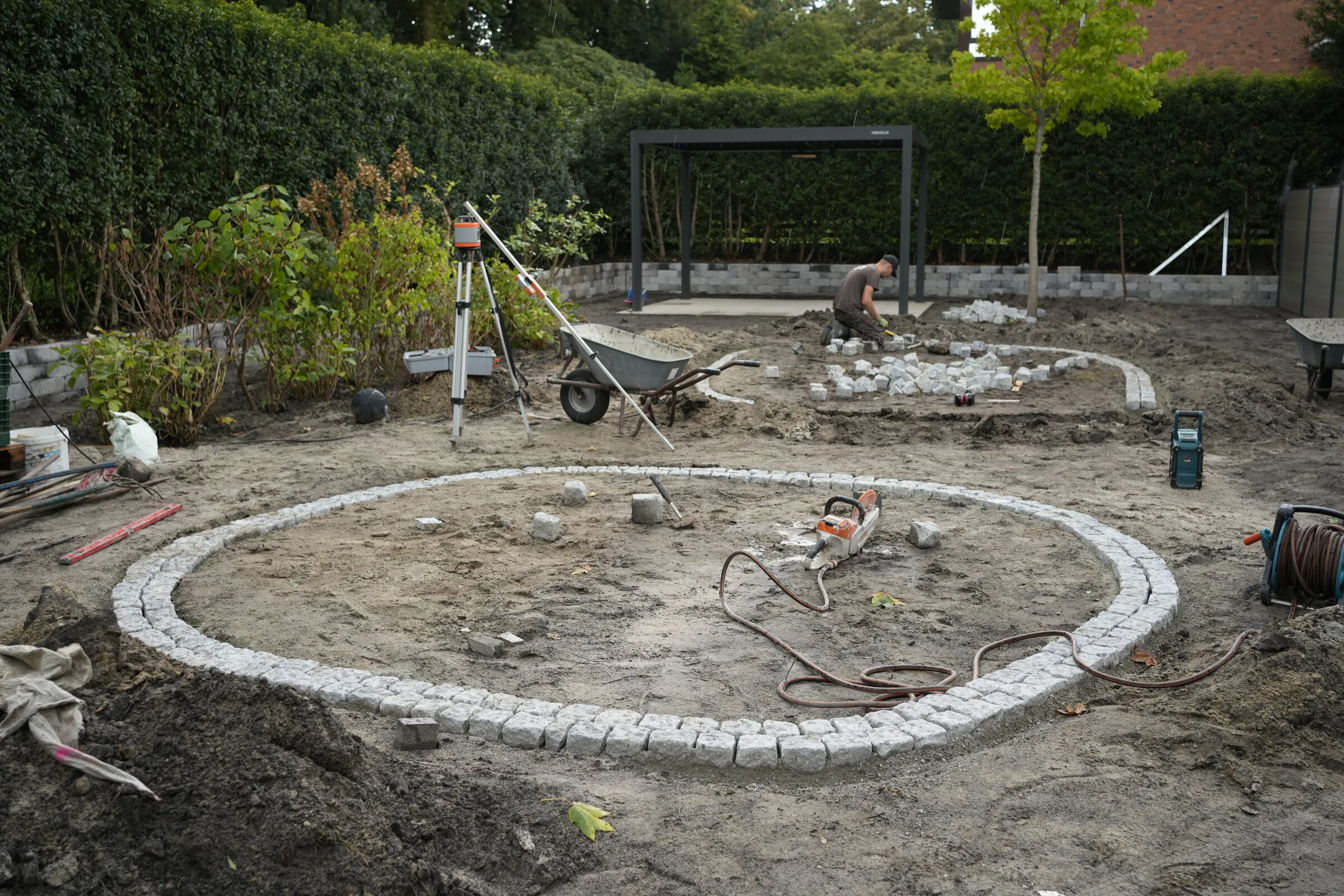Kreisrundes Rondell aus Granitpflaster in Doppelreihe, im Bau, dahinter Rotationslaser auf Stativ und die Pergola